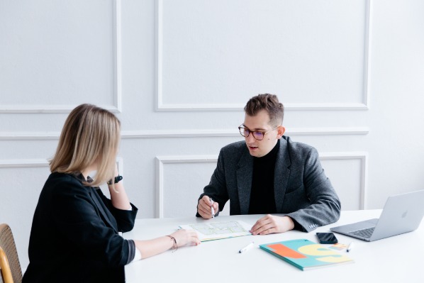 Agent nieruchomości omawia plan mieszkania z klientką przy białym stole, laptop, telefon i notatnik obok.