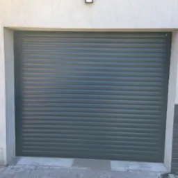 Zamknięta, szara brama garażowa rolowana z widocznym kranem na ścianie obok.