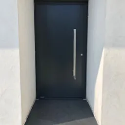 Nowoczesne, antracytowe drzwi wejściowe z długim, pionowym uchwytem, osadzone w białym, surowym murze, z widocznym progiem z ciemnego kamienia.