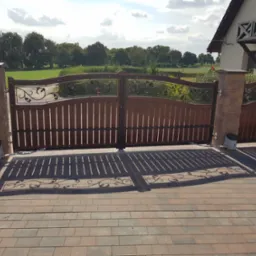 Dwuskrzydłowa brama wjazdowa z drewnianymi panelami i metalowymi zdobieniami, zamontowana na murowanych słupkach z automatyką, rzucająca cień na brukowaną nawierzchnię podjazdu.