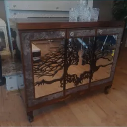 Drewniana komoda z lustrzanymi drzwiczkami, na których wycięto motyw drzewa z profilami twarzy. Blat komody z ciemnego drewna, na nim szklane naczynia. Widoczne dekoracyjne nóżki i otoczenie wnętrza.