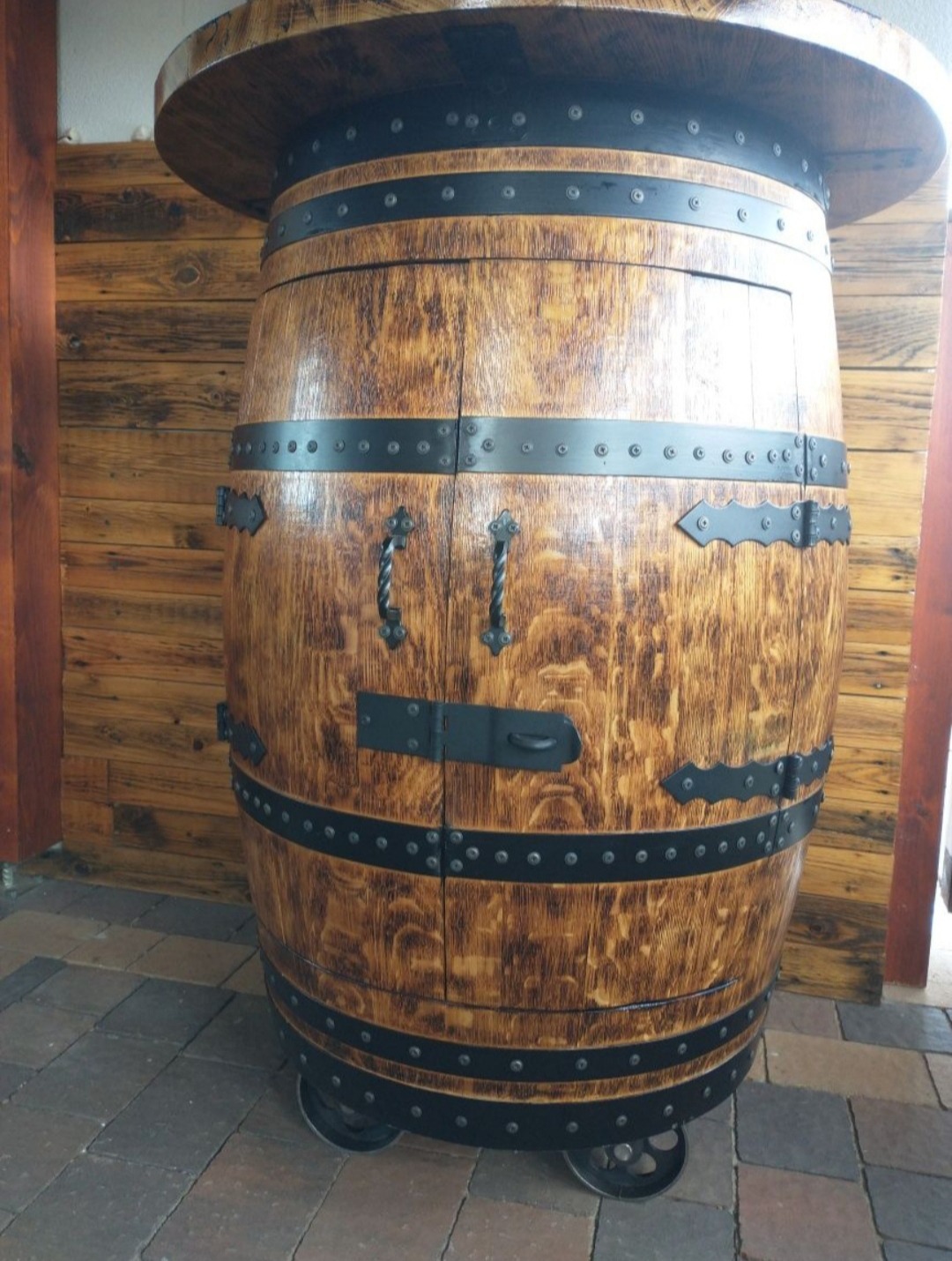 Drewniana beczka z blatem, przerobiona na szafkę z drzwiczkami, wzmocniona czarnymi metalowymi obręczami i elementami dekoracyjnymi, osadzona na kółkach.