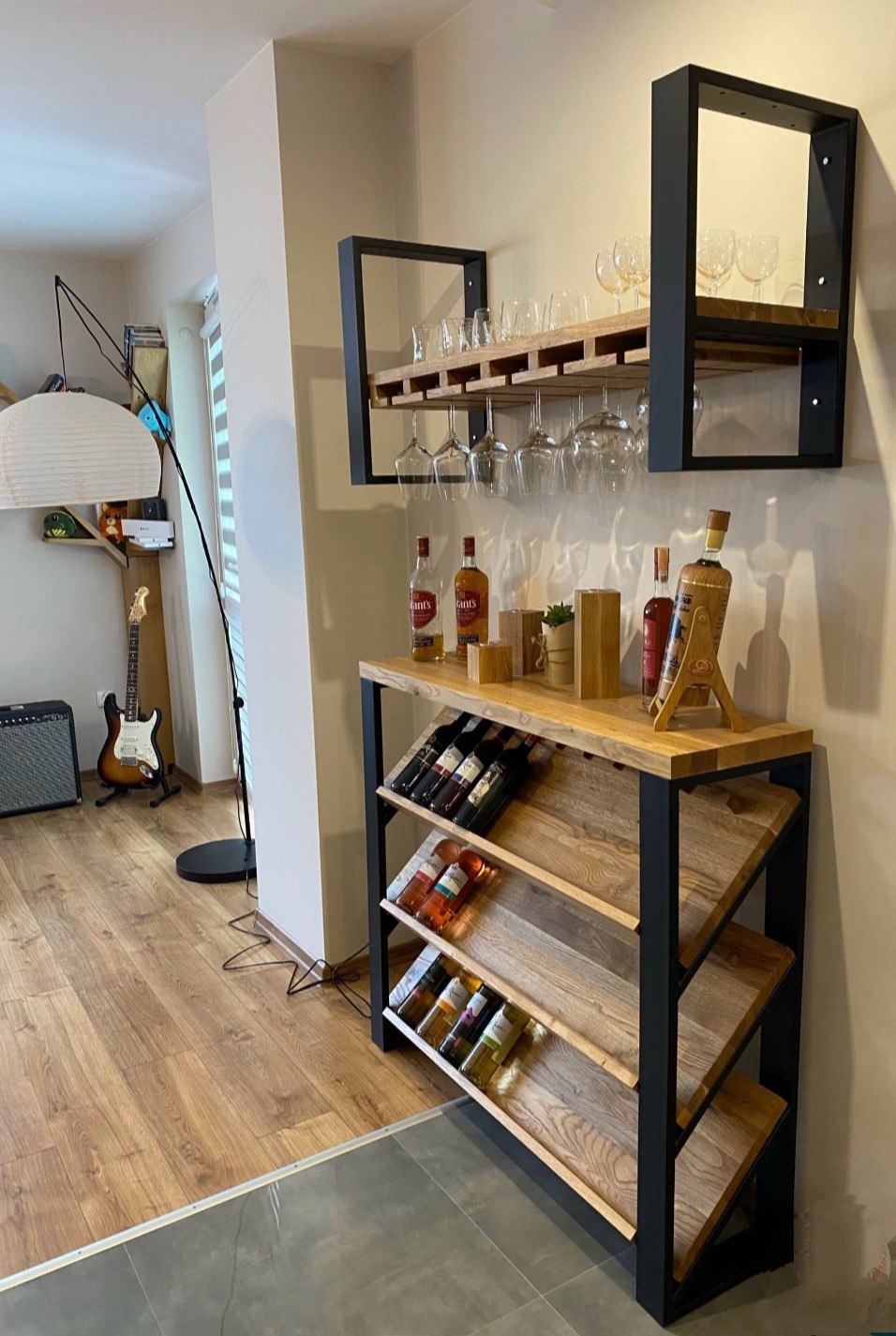 Drewniany barek z czarnymi metalowymi elementami, na blacie butelki alkoholu i dekoracje, nad nim półka na kieliszki i kwadratowa rama, w tle gitara i lampa.