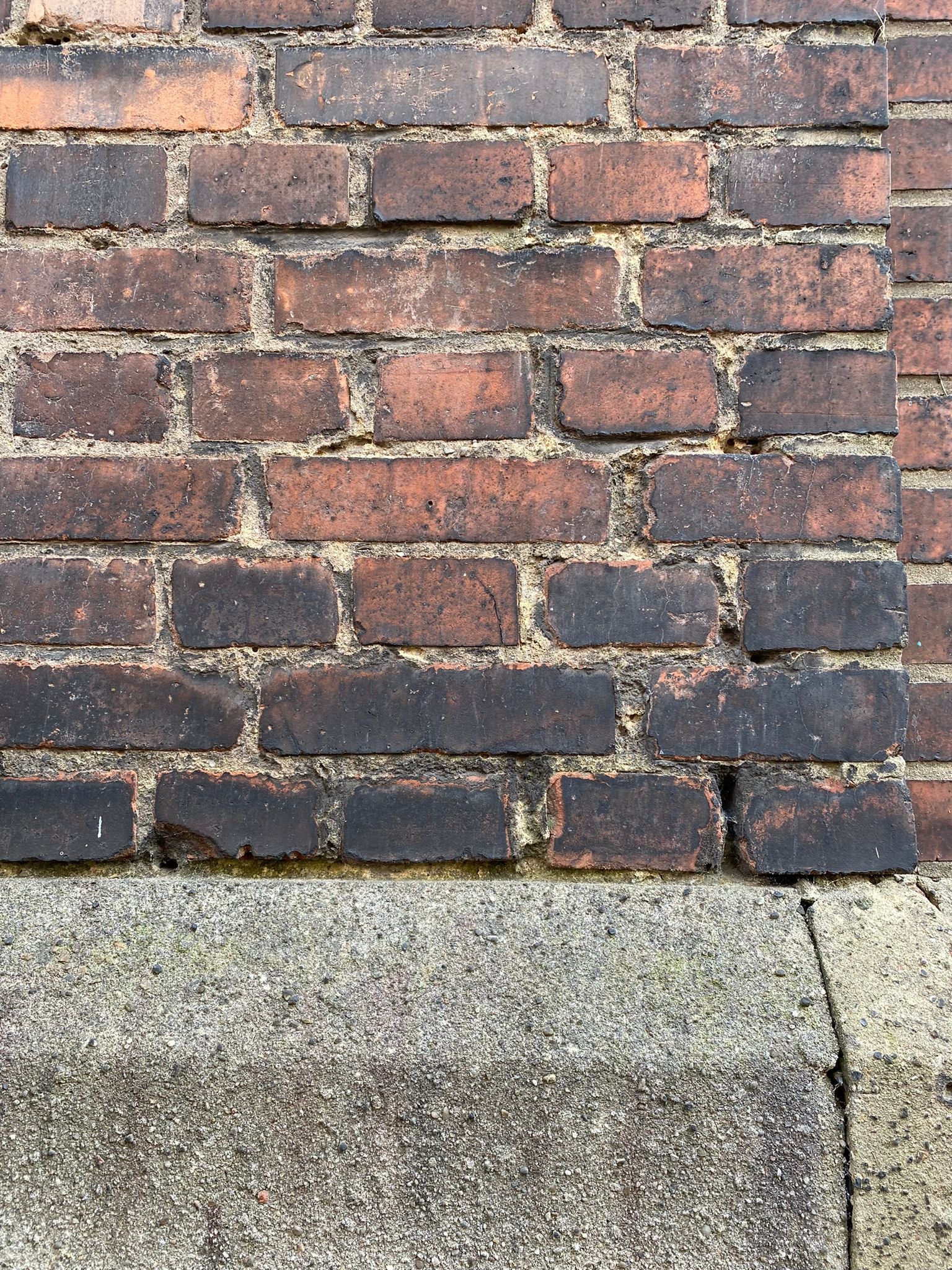 Fragment starej, ciemnobrązowej ściany z cegły klinkierowej, z widocznym spoinowaniem i betonową podmurówką z wyraźną fakturą i pęknięciem w rogu.
