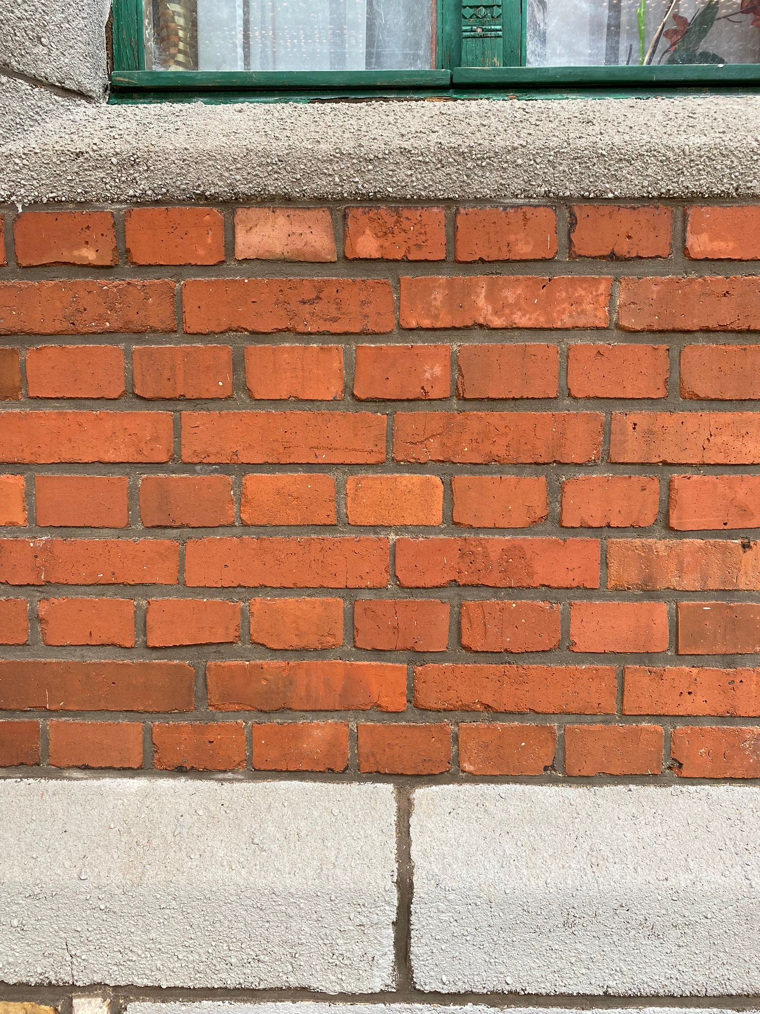 Fragment murowanej ściany z czerwonej cegły z szarą, chropowatą podstawą i górną krawędzią, nad którą widać zielone obramowanie okna.
