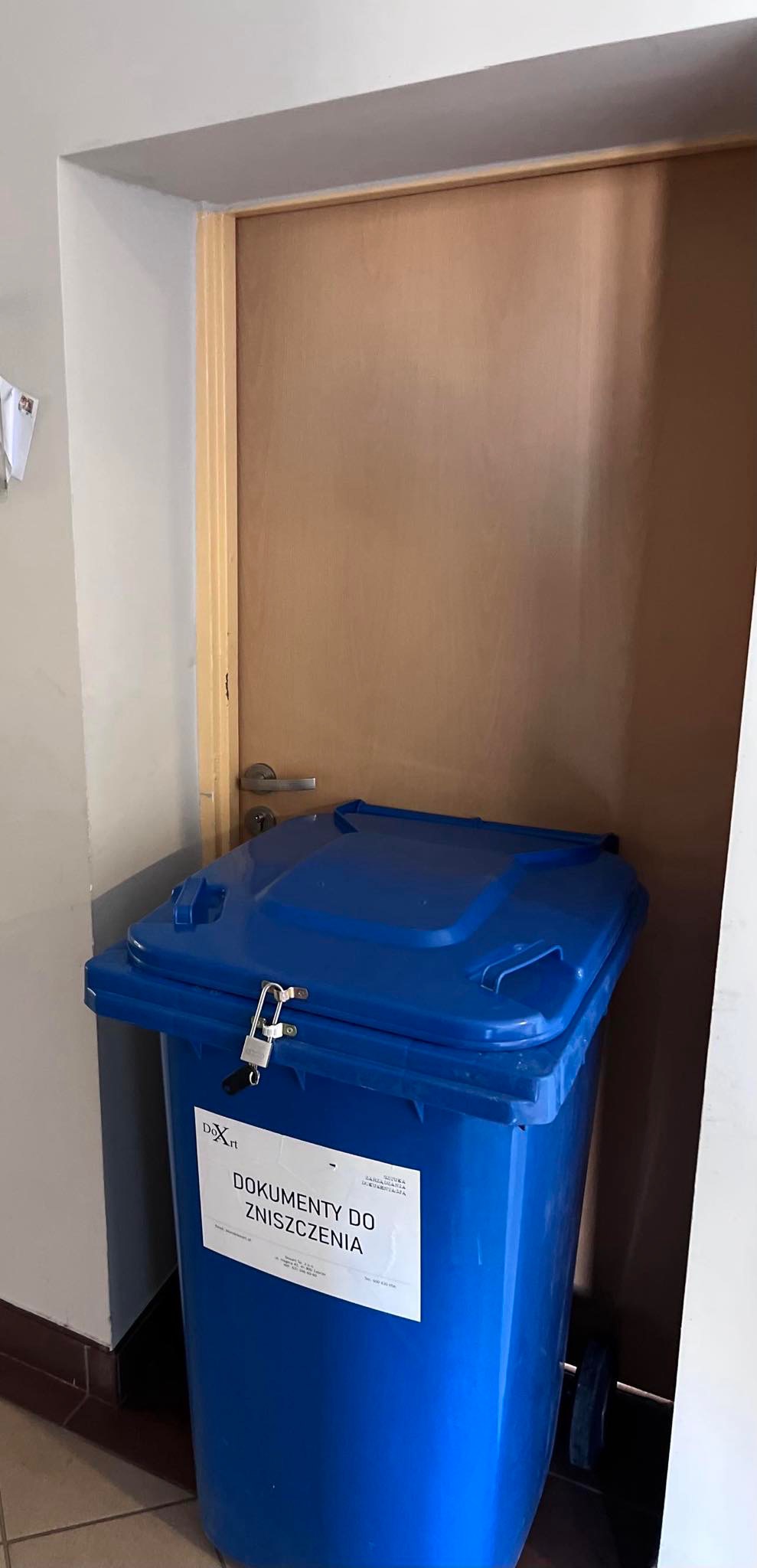 Niebieski, zamykany pojemnik na dokumenty do zniszczenia, z naklejoną etykietą informacyjną, stoi w rogu pomieszczenia przy drzwiach.