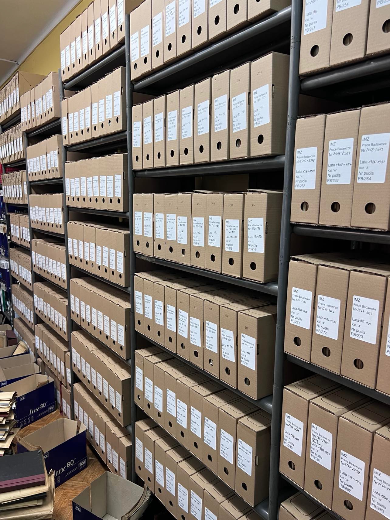 Rzędy tekturowych pudeł archiwizacyjnych na metalowych regałach, wypełnione dokumentami. Widoczne etykiety z opisami zawartości. Perspektywa z dołu, ukazująca głębię magazynu.