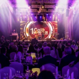 Widok na scenę podczas gali, z zespołem występującym przed dużym ekranem LED, oświetlonym fioletowymi światłami, z widownią siedzącą przy nakrytych stołach.