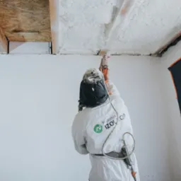 Pracownik w kombinezonie ochronnym aplikuje białą piankę izolacyjną na sufit poddasza z drewnianymi belkami, widoczny sprzęt do natrysku i logo firmy na kombinezonie.