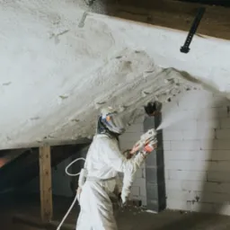 Pracownik w kombinezonie ochronnym aplikuje białą pianę izolacyjną na skośny strop poddasza, widoczne drewniane elementy konstrukcyjne i fragment ściany z bloczków.