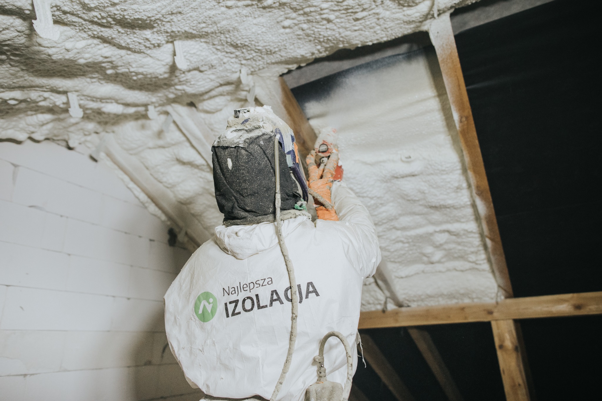 Pracownik w kombinezonie ochronnym z logo firmy 'Najlepsza IZOLACJA' aplikuje piankę izolacyjną na poddaszu, widoczne drewniane elementy konstrukcji dachu.