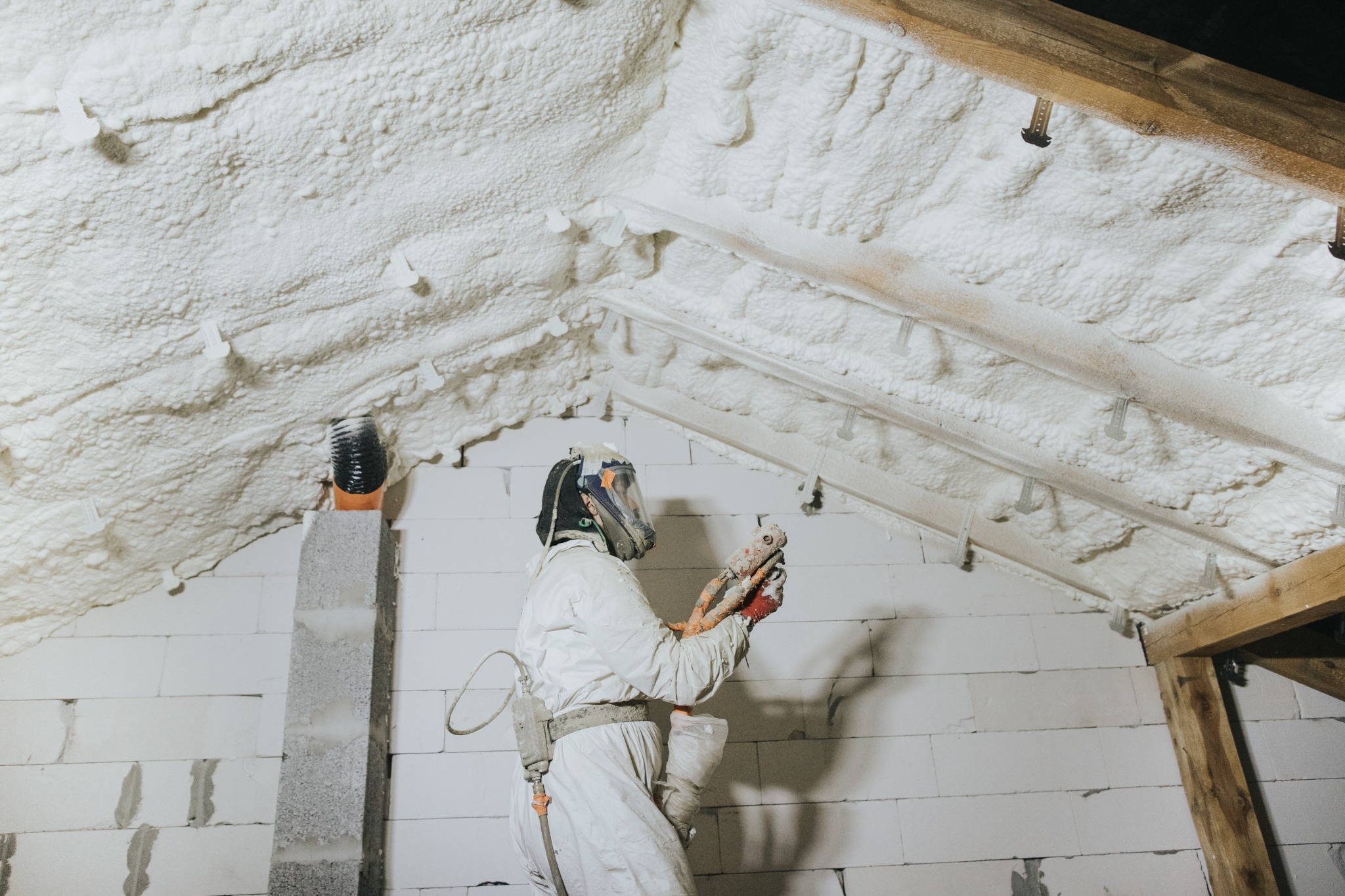 Pracownik w kombinezonie ochronnym aplikuje pianę izolacyjną na poddaszu, widoczne drewniane elementy konstrukcyjne dachu i białe ściany z betonu komórkowego.