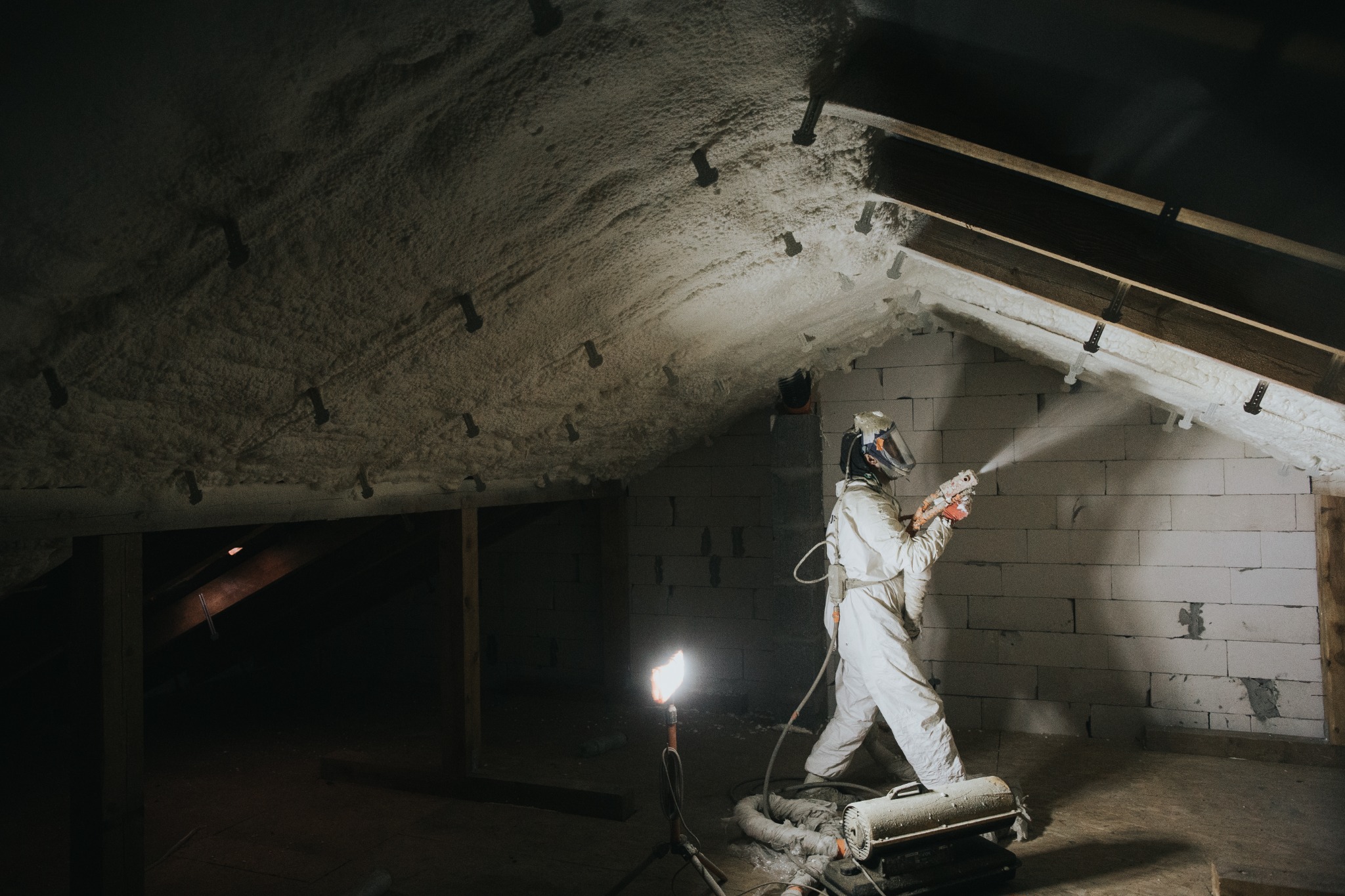 Pracownik w kombinezonie ochronnym aplikuje pianę izolacyjną na poddaszu, widoczne krople piany w powietrzu, oświetlenie halogenowe na statywie, ściany z bloczków betonowych.