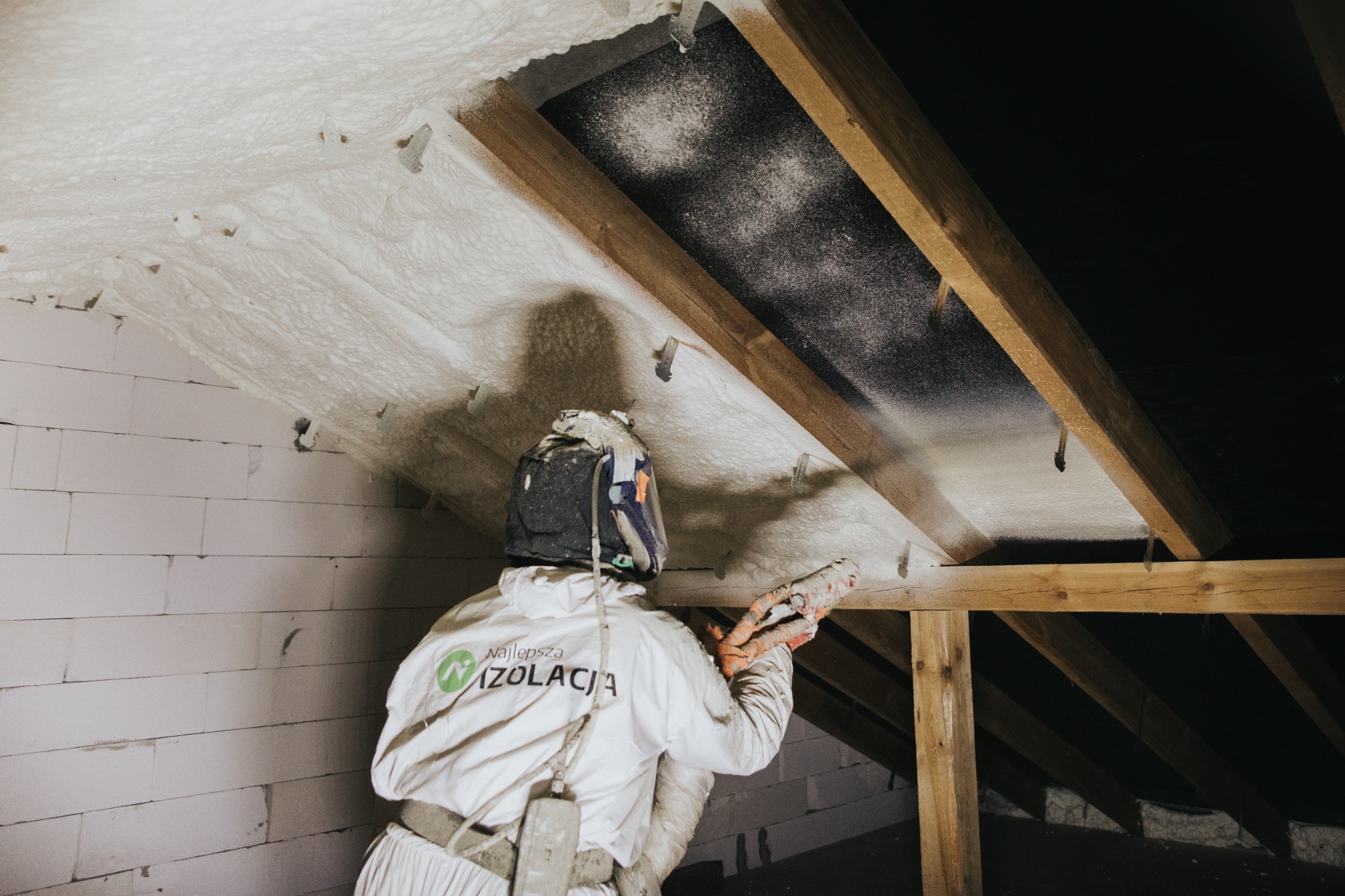 Pracownik w kombinezonie ochronnym aplikuje piankę PUR na poddasze, widoczne krople pianki w powietrzu, drewniana konstrukcja dachu i ściana z bloczków.