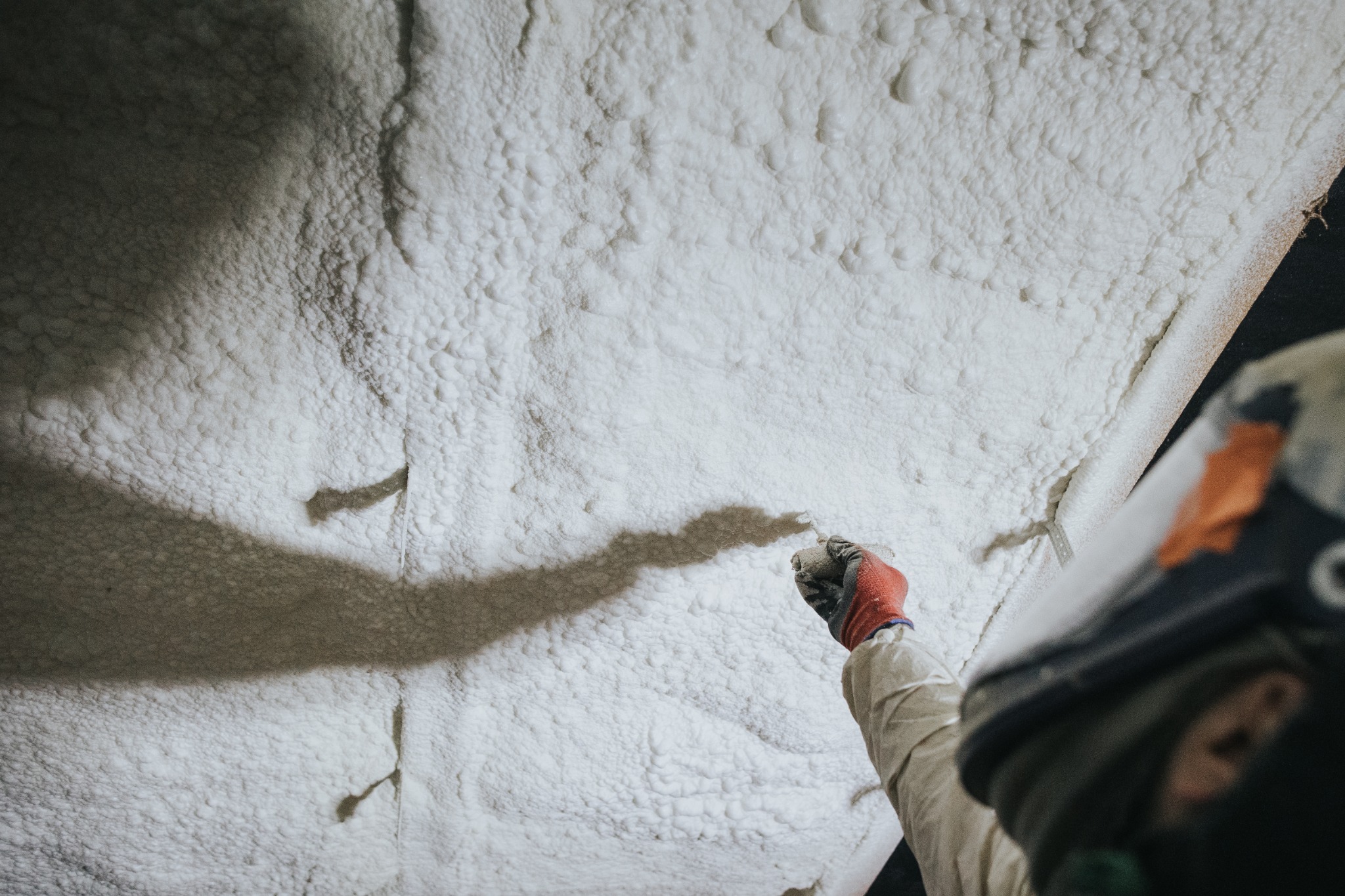 Aplikacja białej piany izolacyjnej na ścianę przez pracownika w kombinezonie i masce ochronnej, widoczny cień ręki i fragment ściany z nałożoną grubą warstwą izolacji.