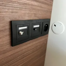 Czarna listwa z gniazdem antenowym, gniazdem RJ45 i gniazdkiem elektrycznym, zamontowana na panelu z drewnopodobną okleiną, obok otworu na kable w białym blacie biurka.