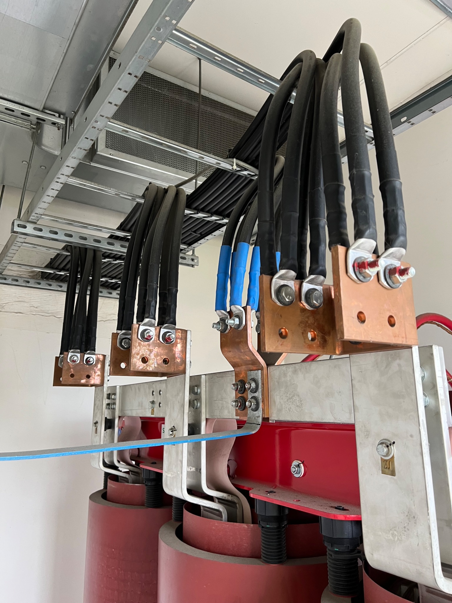 Zestaw grubych kabli elektrycznych w czarnej izolacji, podłączonych do miedzianych złączek i szyn zbiorczych, zamocowanych na metalowej konstrukcji wewnątrz rozdzielni.