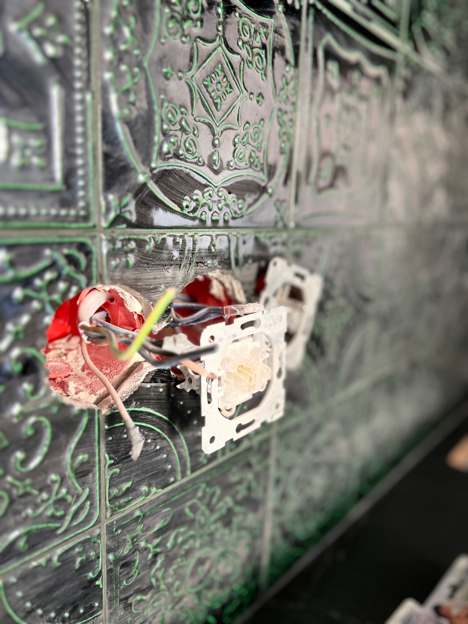 Montaż gniazdka elektrycznego w ozdobnych, ciemnych płytkach ściennych z zielonymi wzorami, widoczne przewody i elementy mocujące.