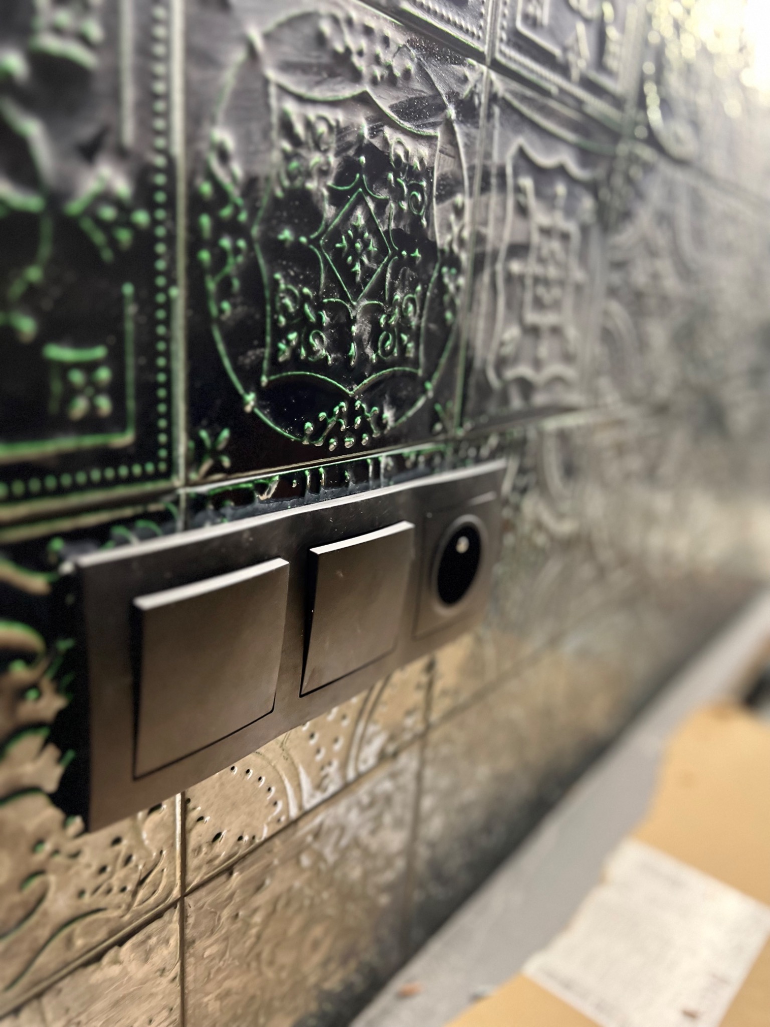 Dwa czarne, matowe włączniki światła i gniazdko elektryczne zamontowane na dekoracyjnej, tłoczonej ścianie w stylu retro z zielonymi akcentami.