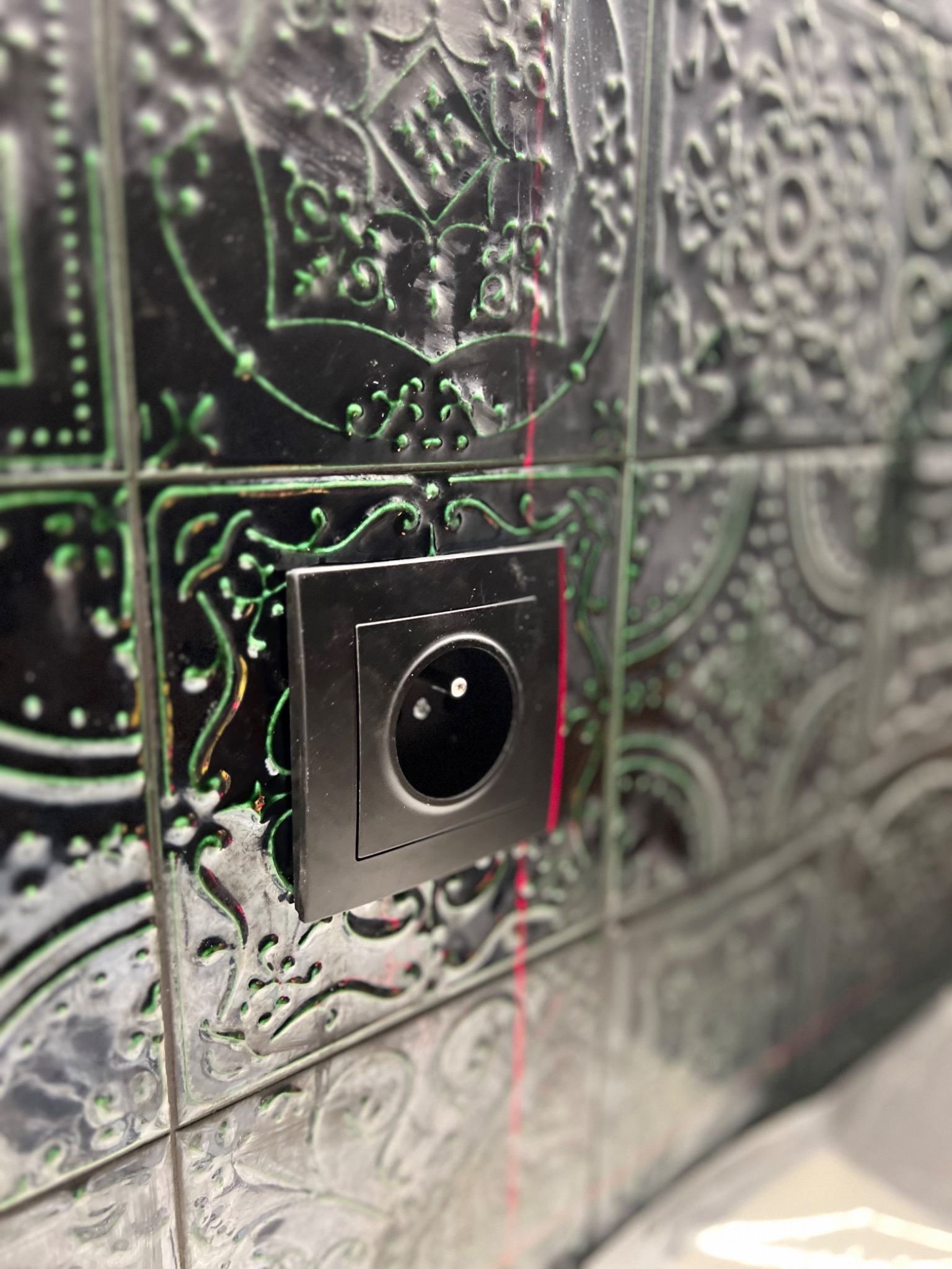 Czarny kontakt elektryczny zamontowany na ozdobnych, czarno-zielonych płytkach ściennych z wytłaczanym wzorem, oświetlony delikatnym czerwonym światłem lasera.