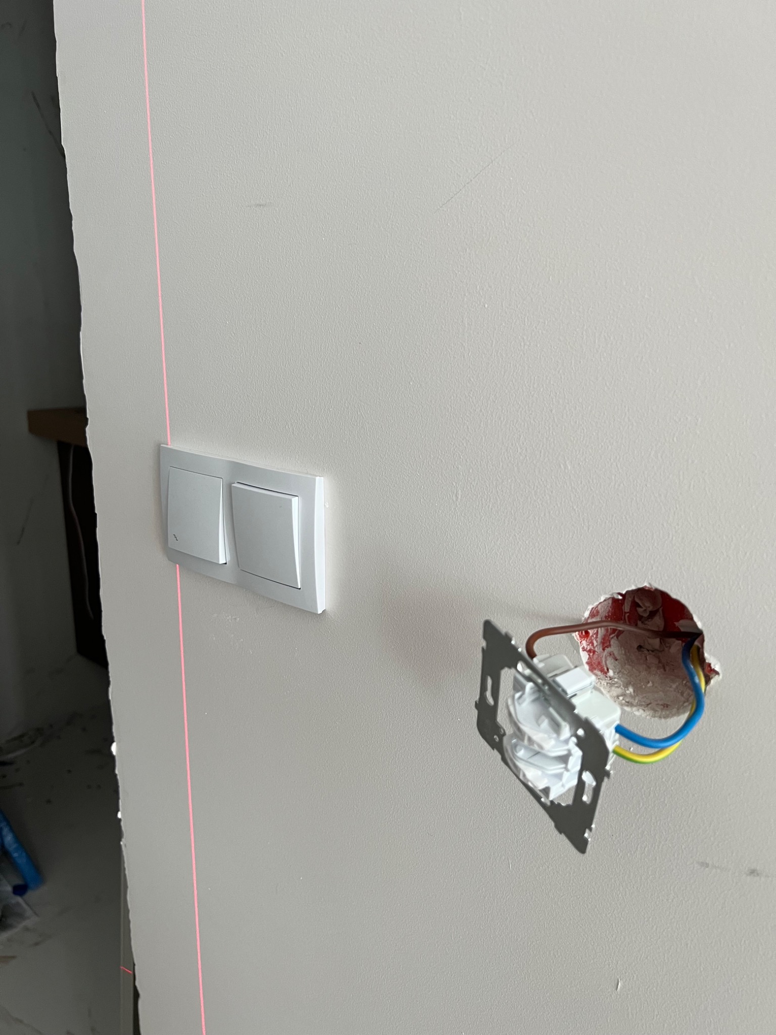 Montaż podwójnego włącznika światła na szarej ścianie, widoczna wiązka lasera wyznaczająca pion, obok puszka elektryczna z wystającymi przewodami.