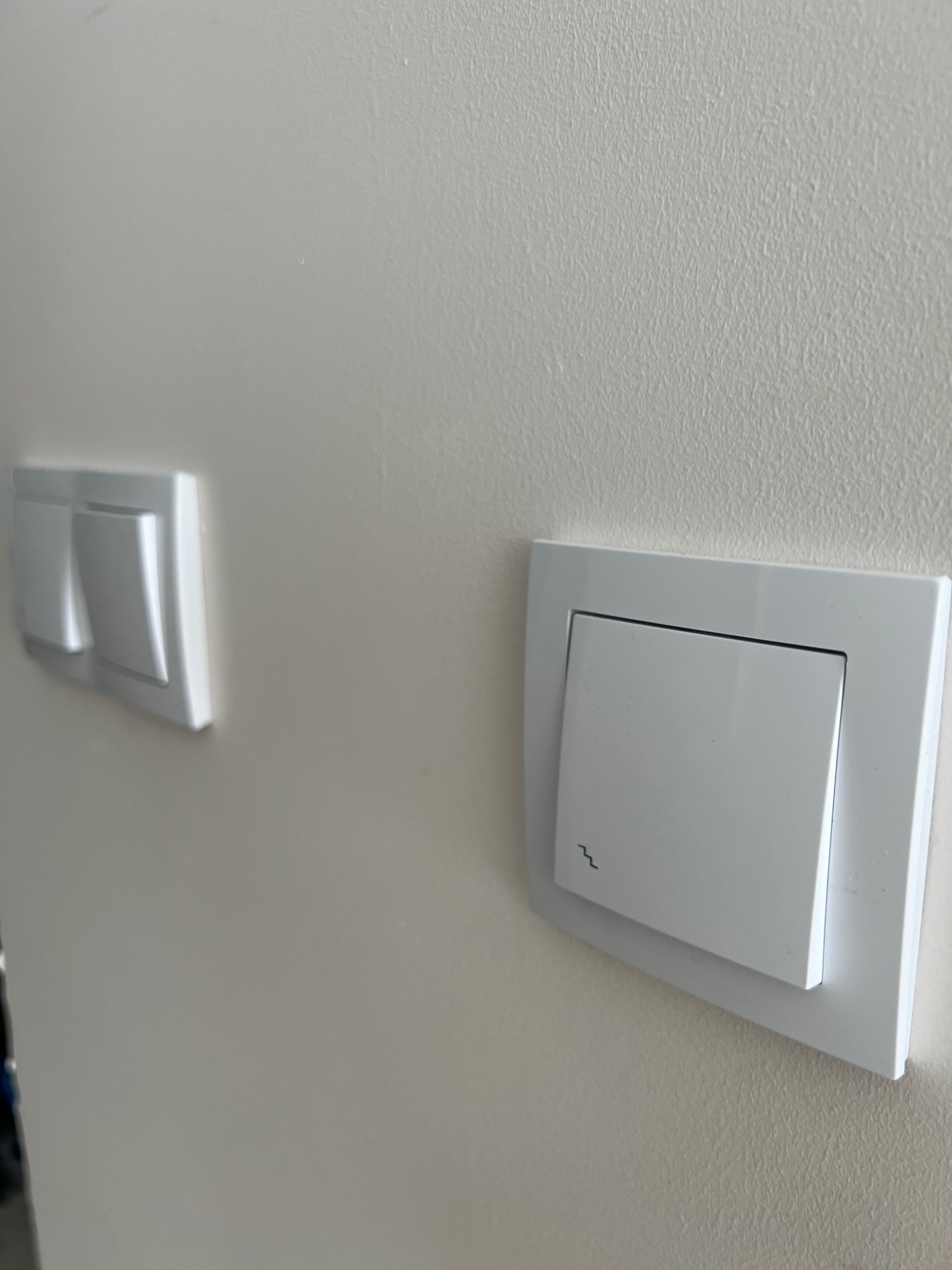 Dwa białe, nowoczesne włączniki światła zamontowane na jasnej, kremowej ścianie. Jeden włącznik z symbolem fali na dolnej części klawisza.