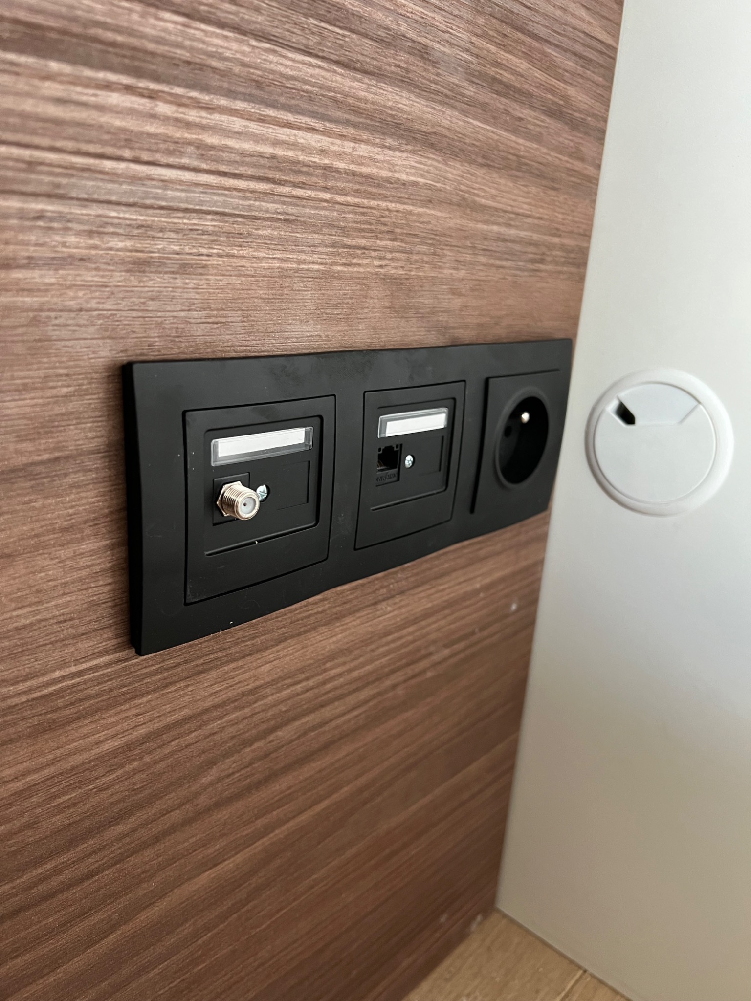 Czarna listwa z gniazdem antenowym, gniazdem RJ45 i gniazdkiem elektrycznym, zamontowana na panelu z drewnopodobną okleiną, obok otworu na kable w białym blacie biurka.