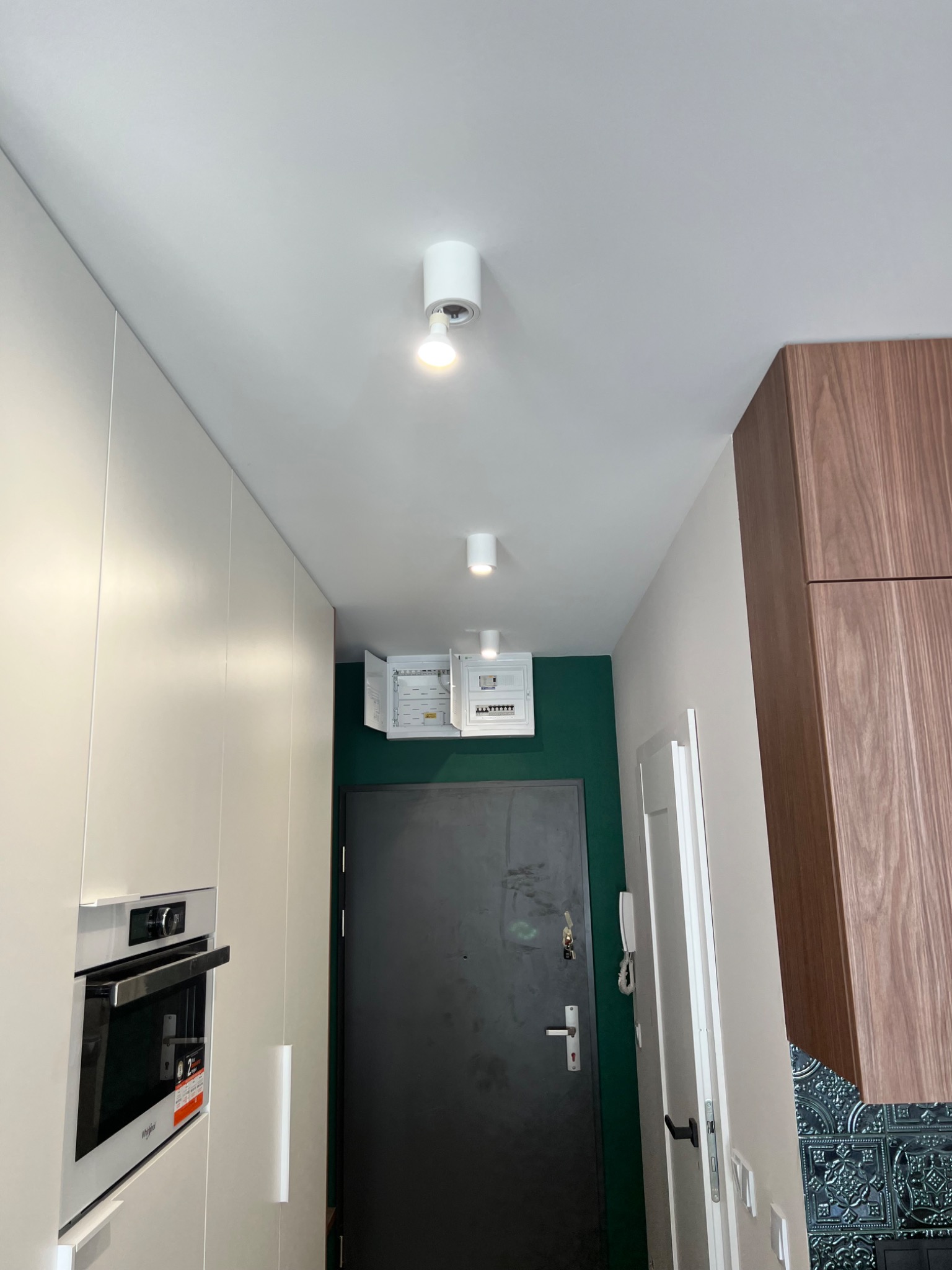 Wnętrze mieszkania z otwartą skrzynką bezpiecznikową na ścianie pomalowanej na zielono, widoczne białe szafki kuchenne, ciemne drzwi wejściowe i dwa białe reflektory sufitowe.