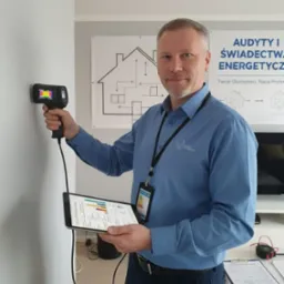 Mężczyzna z kamerą termowizyjną bada ścianę, trzymając tablet z danymi. W tle plakat o audycie energetycznym i świadectwach.