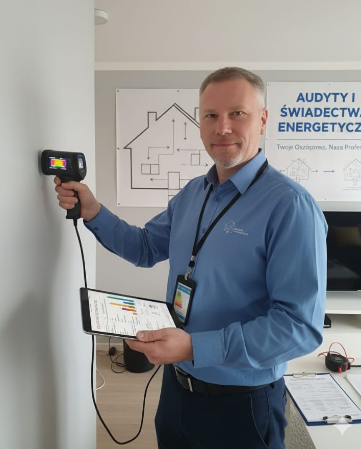 Mężczyzna z kamerą termowizyjną bada ścianę, trzymając tablet z danymi. W tle plakat o audycie energetycznym i świadectwach.