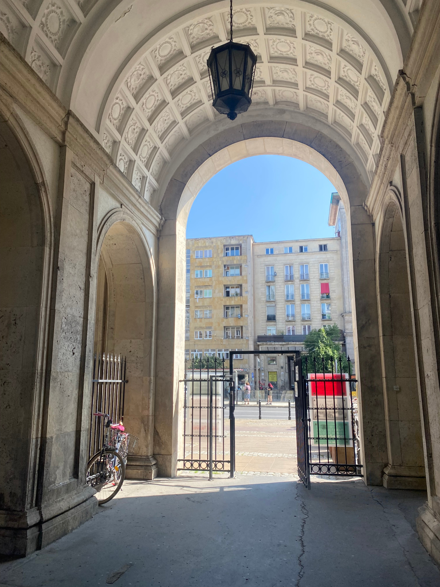 Arched stone entryway with ornate ceiling and hanging lantern, leading to a city street with buildings and pedestrians, a bicycle parked to the side.