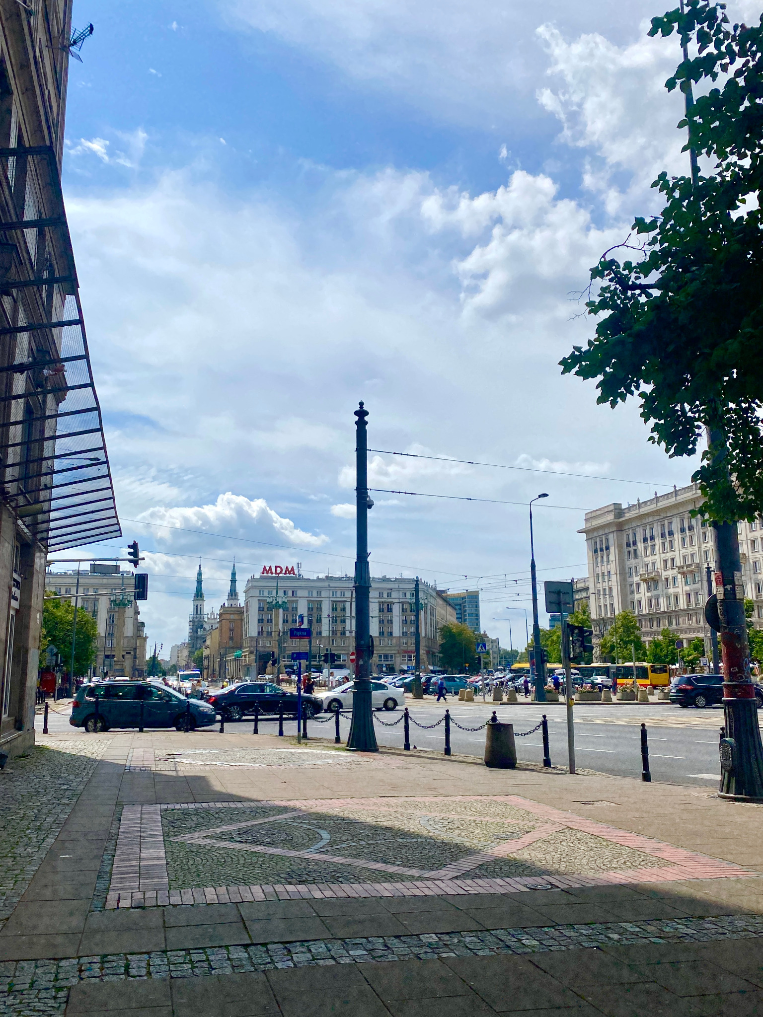 Ulica w centrum miasta z widokiem na budynki w stylu socrealizmu, chodnik z dekoracyjną kostką, ruch uliczny i błękitne niebo z chmurami w tle.