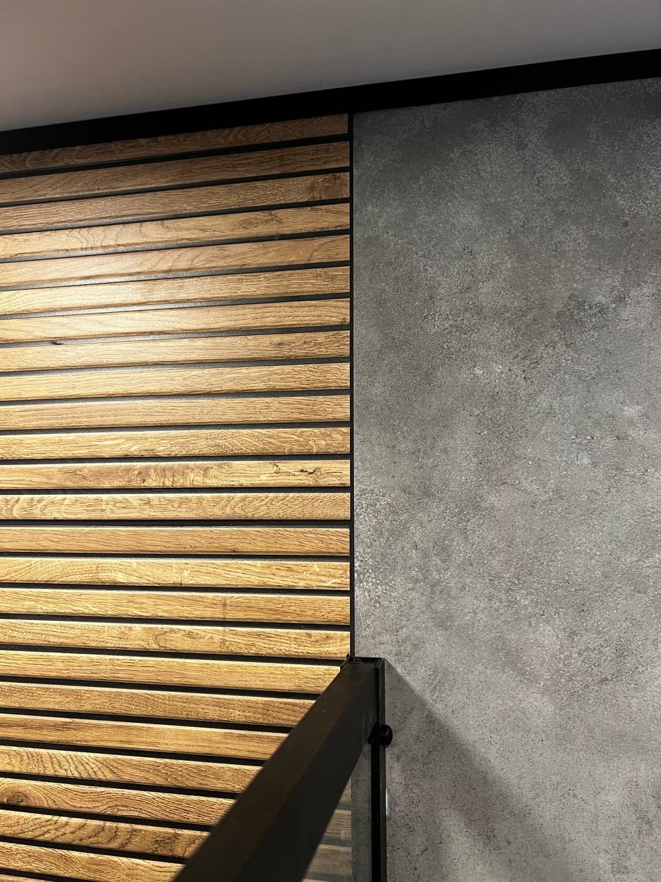 Drewniane lamele ścienne w ciepłym odcieniu drewna sąsiadują z szarą, betonową ścianą. Czarny profil wyznacza górną krawędź kompozycji.