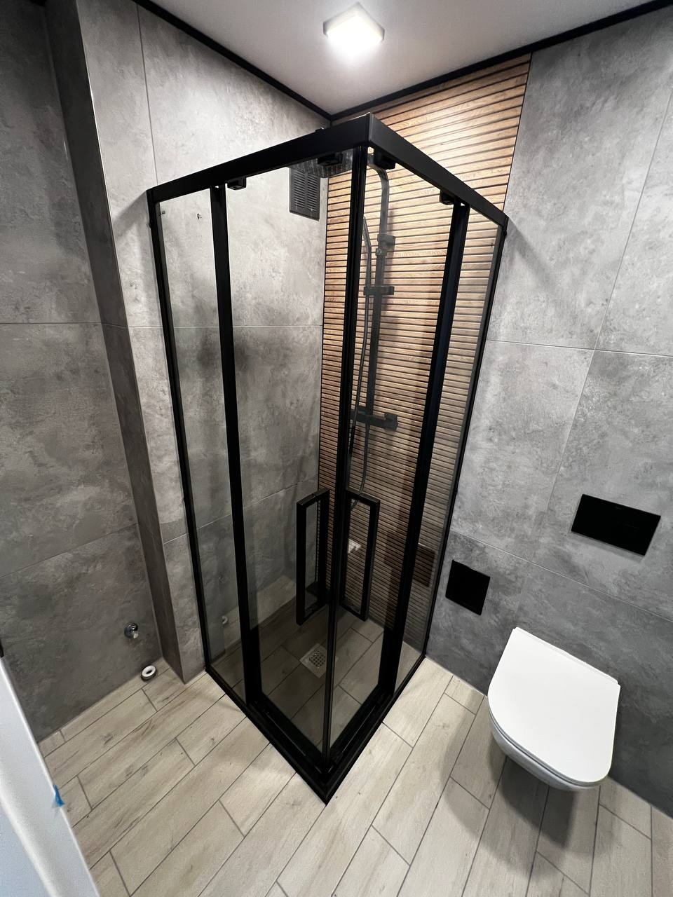 Nowoczesna kabina prysznicowa z czarnymi profilami i drewnianą ścianą w tle, uzupełniona szarą płytką i podwieszaną toaletą.