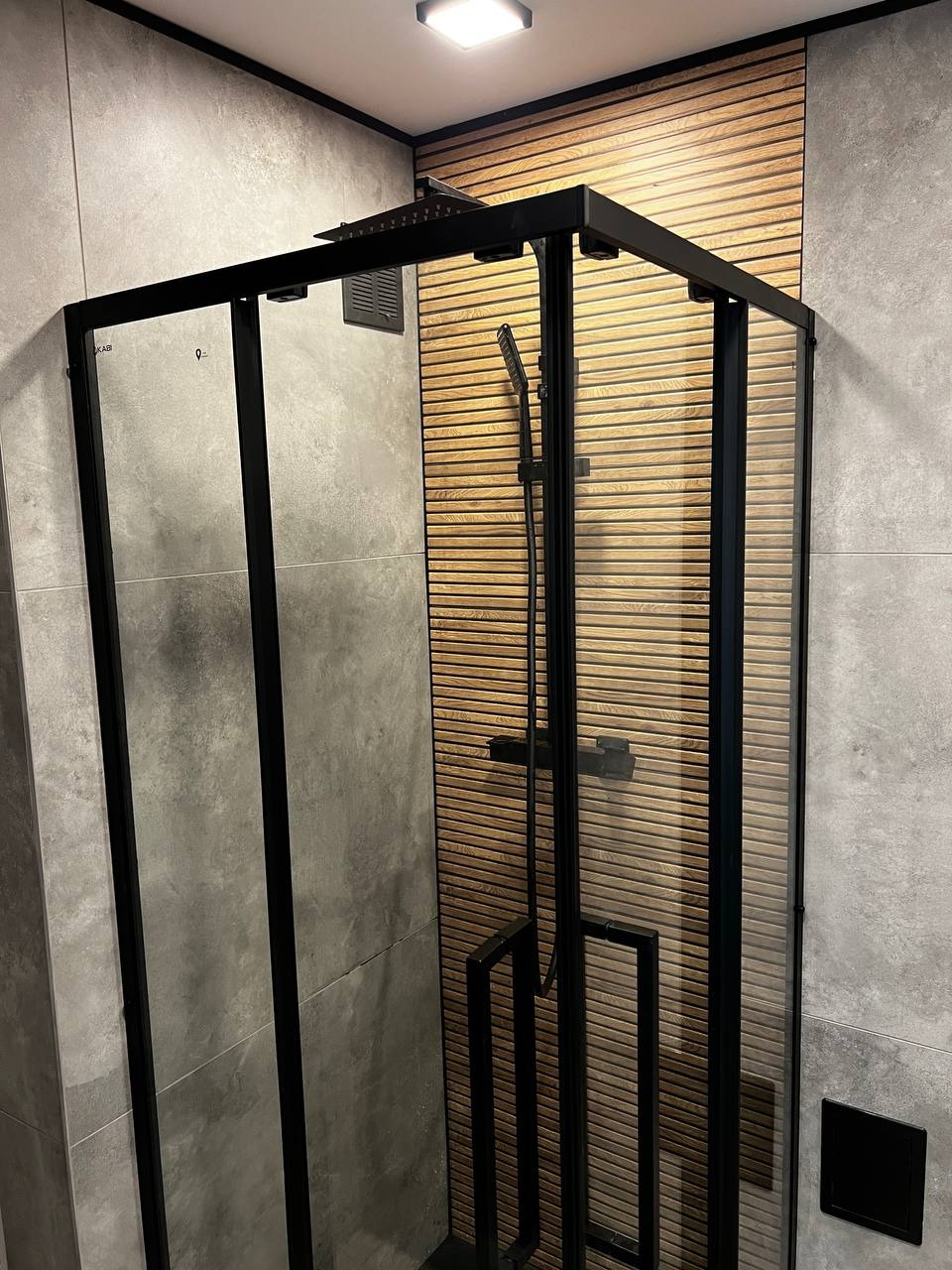 Nowoczesna kabina prysznicowa z czarnymi profilami na tle szarych płytek i drewnianej ściany z lameli. Minimalistyczny design łazienki po remoncie.