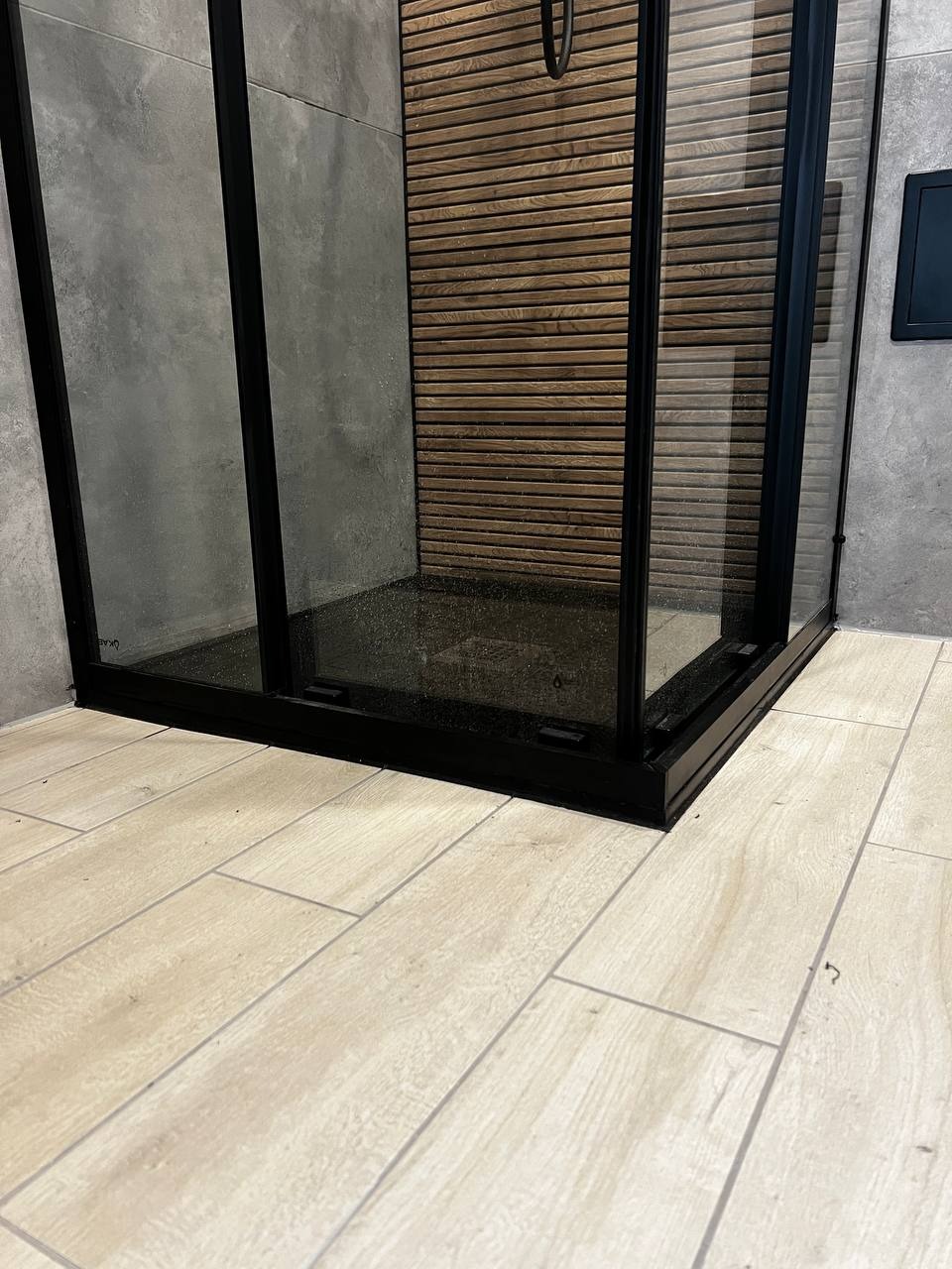 Nowoczesna kabina prysznicowa z czarnymi ramami, drewnianą ścianą i beżowymi płytkami podłogowymi. Minimalistyczny design, eleganckie wykończenie łazienki.