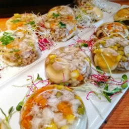 Galaretki z mięsem i warzywami ułożone na białych talerzach, udekorowane świeżymi kiełkami i ziołami, na drewnianym blacie.