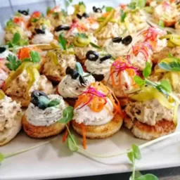 Elegancko ułożone kanapeczki na białym talerzu, z różnymi pastami, oliwkami, ogórkiem konserwowym, marchewką i mikro-ziołami, tworzące apetyczną kompozycję.
