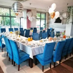 Uroczysta aranżacja stołu na przyjęcie: długi stół nakryty białym obrusem, niebieskie krzesła z weluru, srebrne lampy kuliste, dekoracje kwiatowe i balony w tonacji bieli i złota.