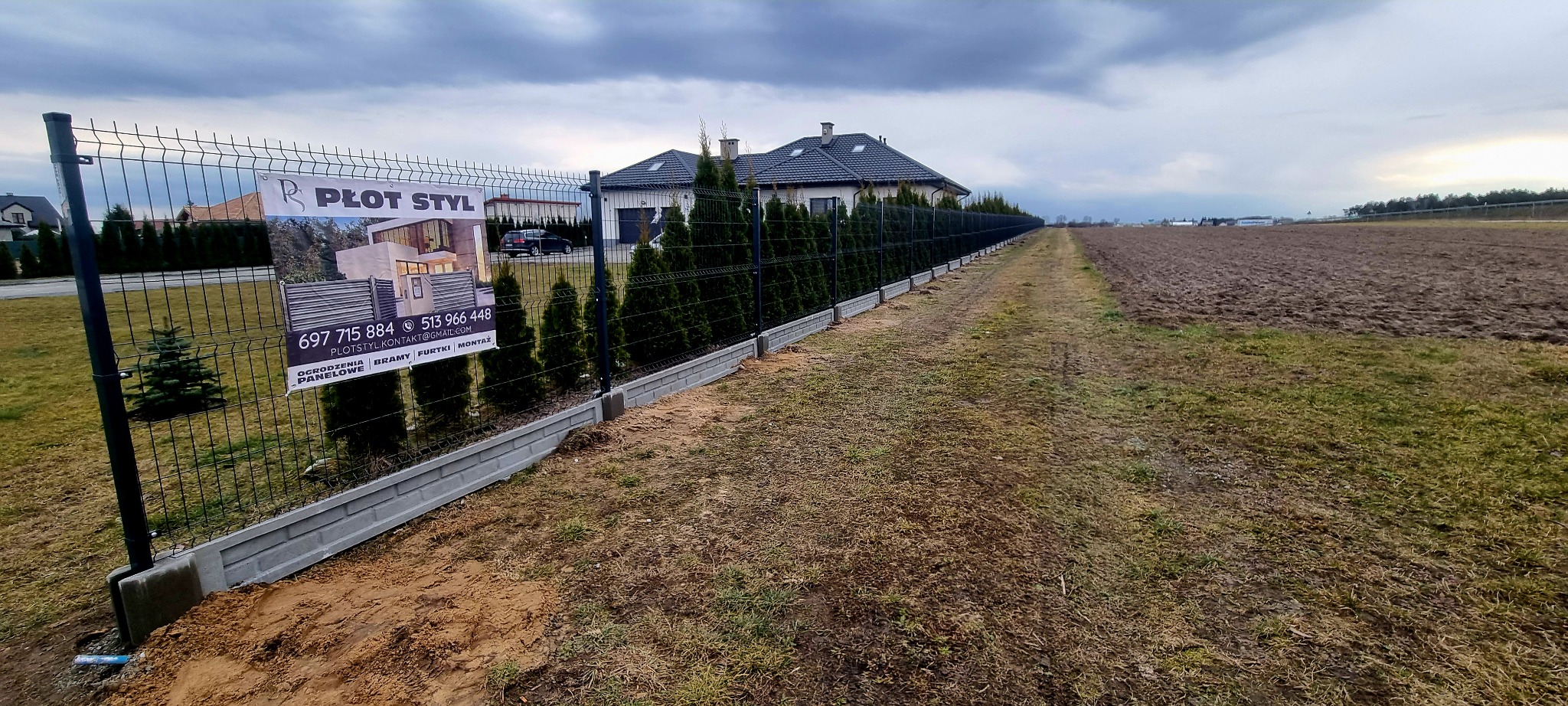 Długa linia ciemnego ogrodzenia panelowego z podmurówką betonową oddzielająca posesję od pola, z widocznym banerem reklamowym firmy 'Płot Styl' i rzędem tuj wzdłuż ogrodzenia.