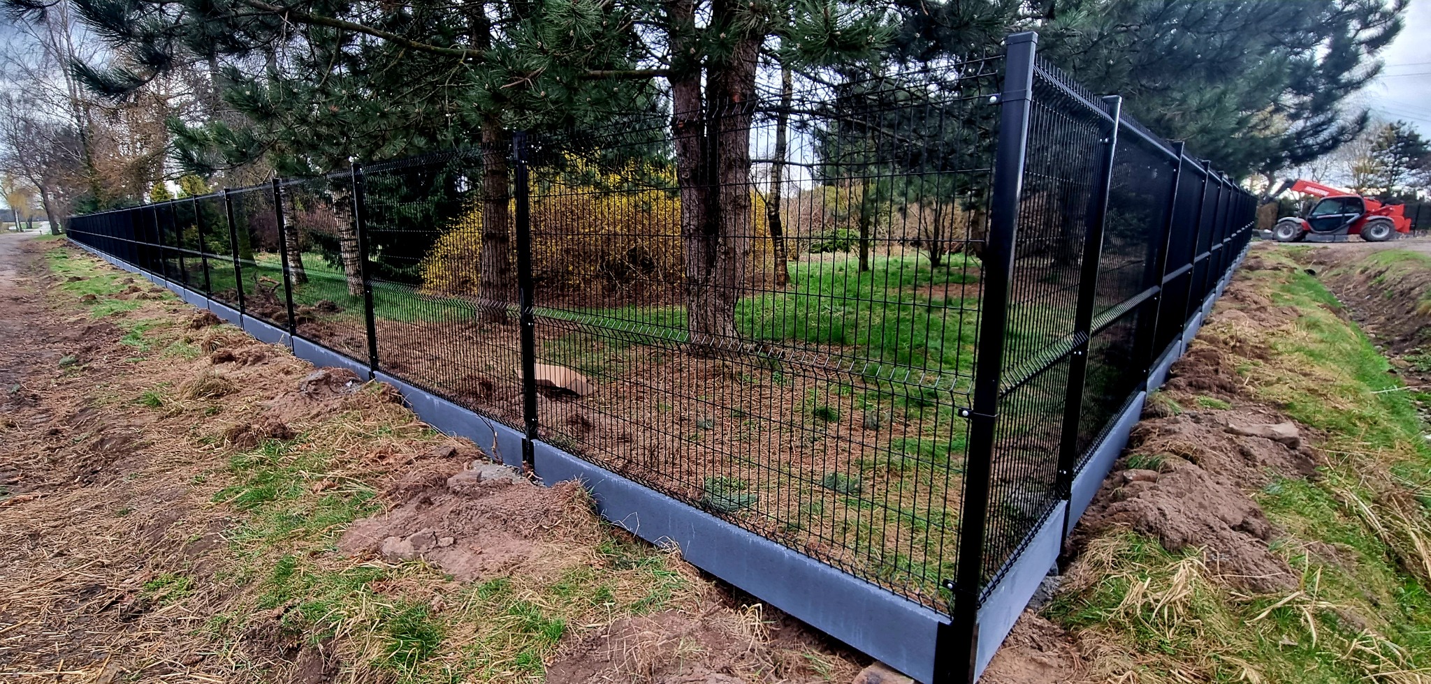 Czarny, systemowy płot z siatki panelowej na betonowej podmurówce, widok z perspektywy gruntu, w tle drzewa i czerwona ładowarka teleskopowa.