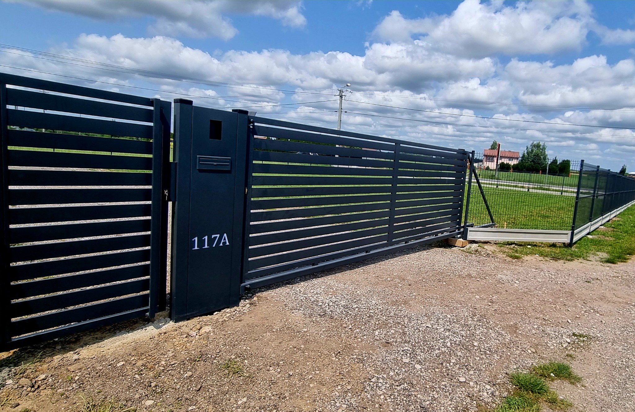 Nowoczesna, przesuwna brama wjazdowa z poziomymi panelami w kolorze antracytowym, z numerem domu 117A i skrzynką na listy, na tle zielonego trawnika i siatki ogrodzeniowej.