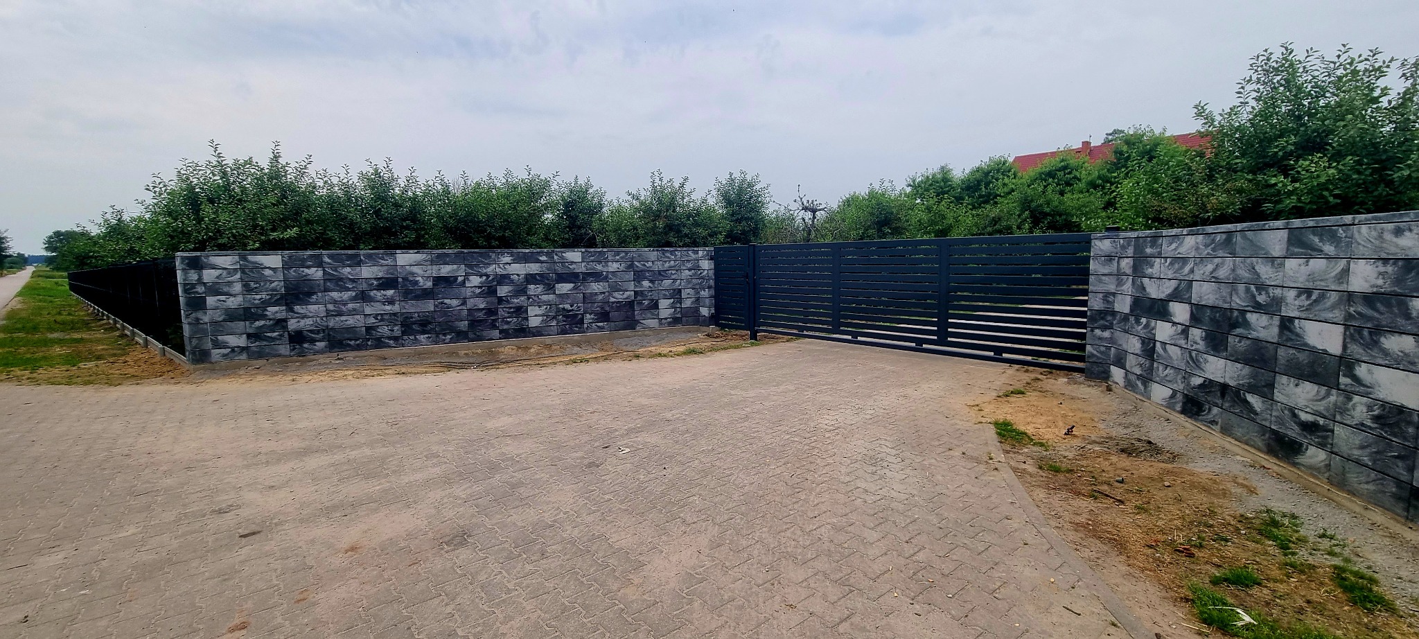 Nowoczesne ogrodzenie z szarej kostki betonowej z poziomą bramą wjazdową na posesję, widok z zewnątrz.