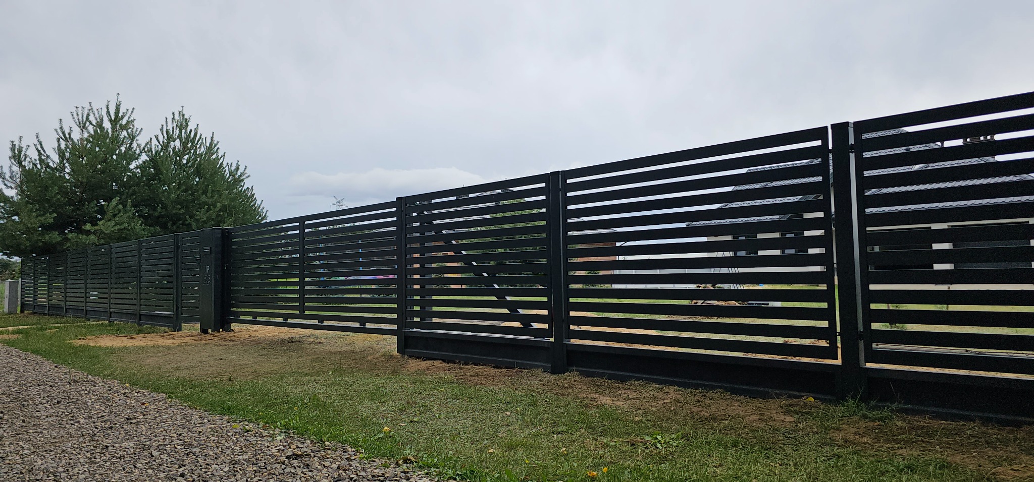 Nowoczesna, pozioma brama przesuwna z czarnych paneli, widoczna od strony zewnętrznej posesji, z fragmentem ogrodzenia panelowego w tle i zielenią trawnika u dołu.