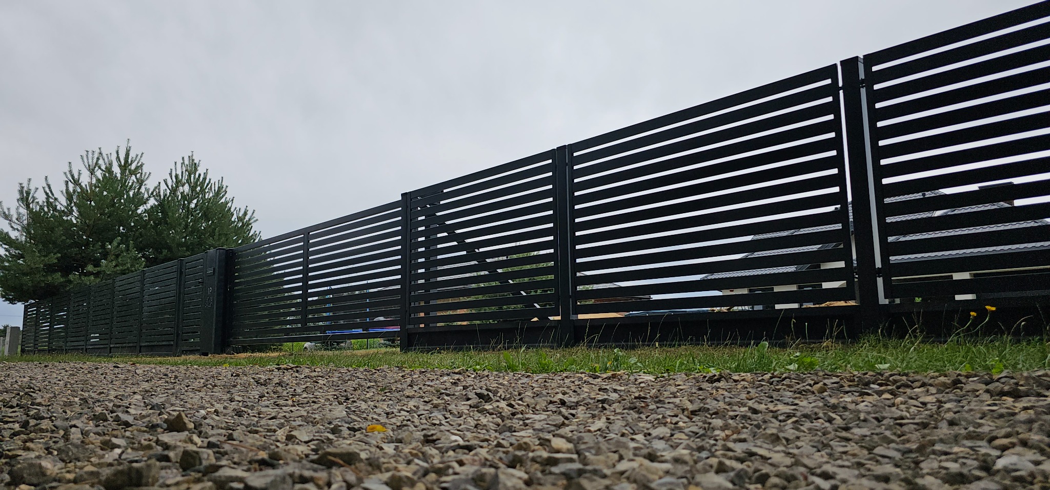 Nowoczesne, czarne ogrodzenie panelowe z poziomymi szczeblami, widok z poziomu gruntu na żwirowej ścieżce, w tle zieleń i fragment budynku.