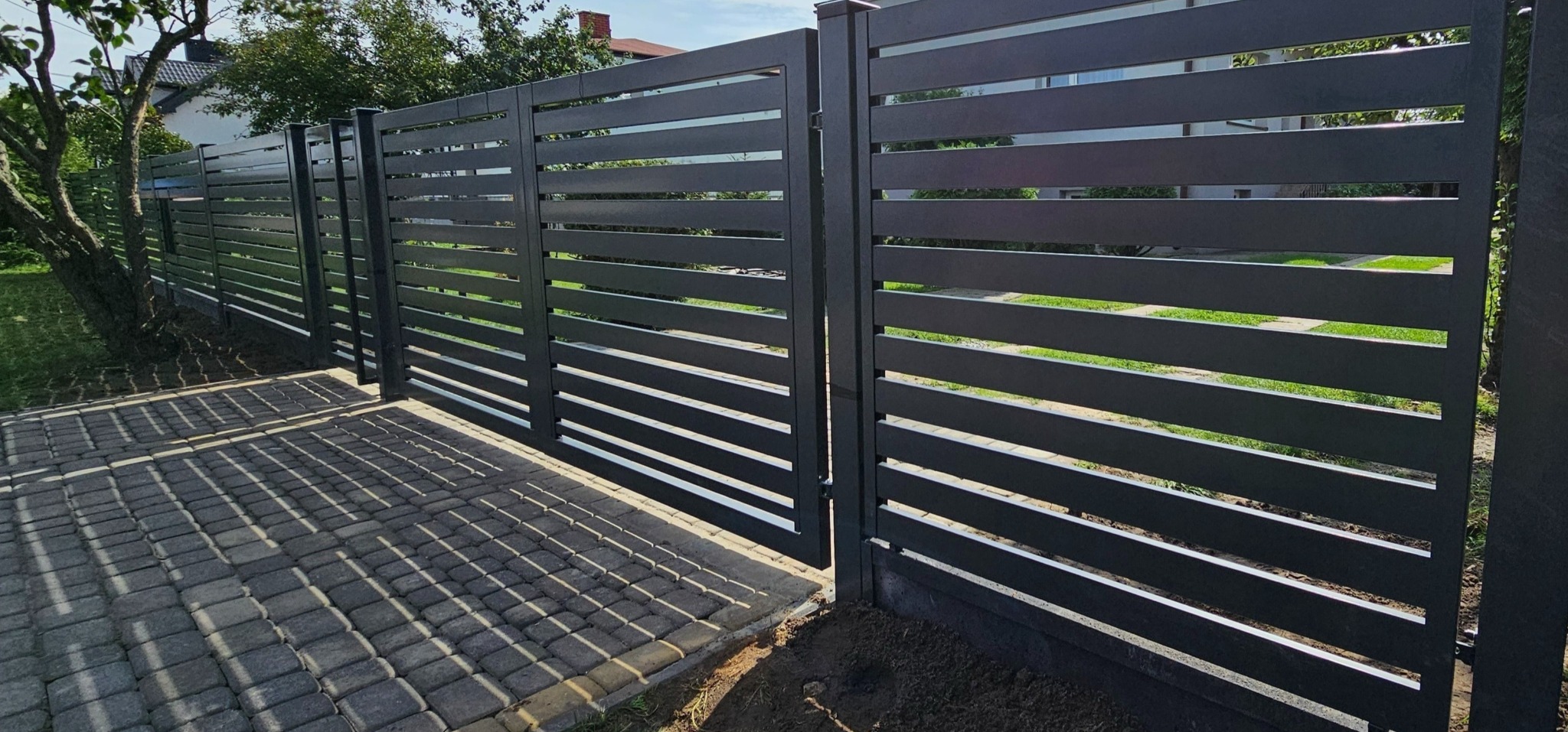 Nowoczesne, poziome, szare ogrodzenie z paneli z bramą wjazdową, widoczne z zewnątrz posesji, z kostką brukową na podjeździe.