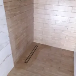 Ukończona kabina prysznicowa z odpływem liniowym, wyłożona płytkami imitującymi drewno, z białymi płytkami marmurkowymi po lewej stronie.