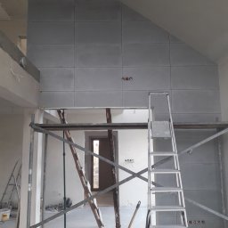 Dariusz Kowalczyk - Wnętrze w trakcie prac wykończeniowych: ściana wyłożona szarymi płytami, widoczne rusztowanie i drabina, przygotowane gniazdka elektryczne.