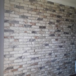 Dariusz Kowalczyk - Ściana z dekoracyjnej cegły w odcieniach bieli, szarości i brązu, z ciemną drewnianą podłogą u dołu kadru.