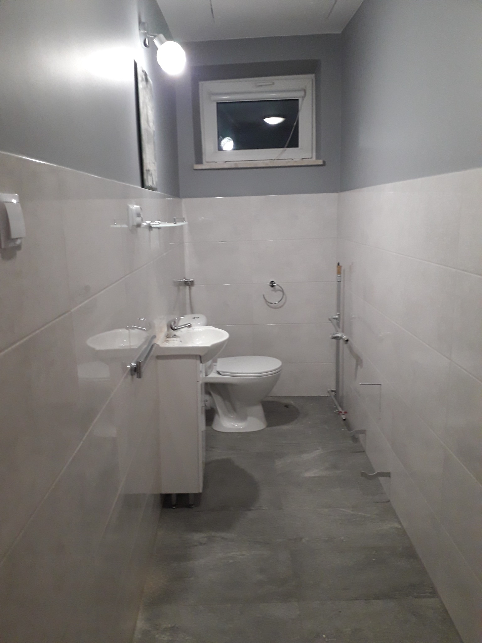 Ukończona łazienka z białymi płytkami na ścianach, szarą górną częścią ścian, umywalką z szafką, toaletą i nową instalacją wodną.